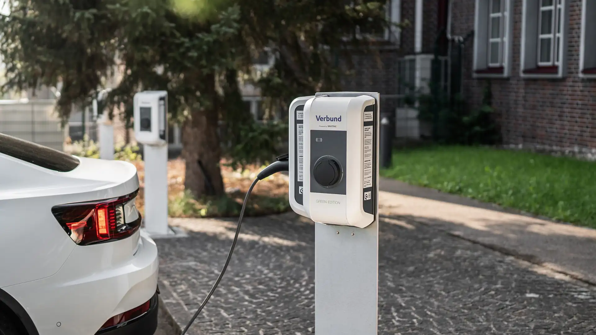 An electric vehicle is being charged at a VERBUND charging station in the parking lot of a company, with multiple charging points and trees nearby.